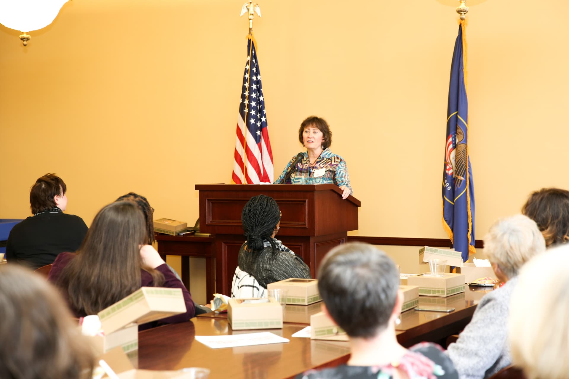 Dr. Cathy Callow-Heusser speaking at a podium before a seated audience, with the American and Utah flags behind her